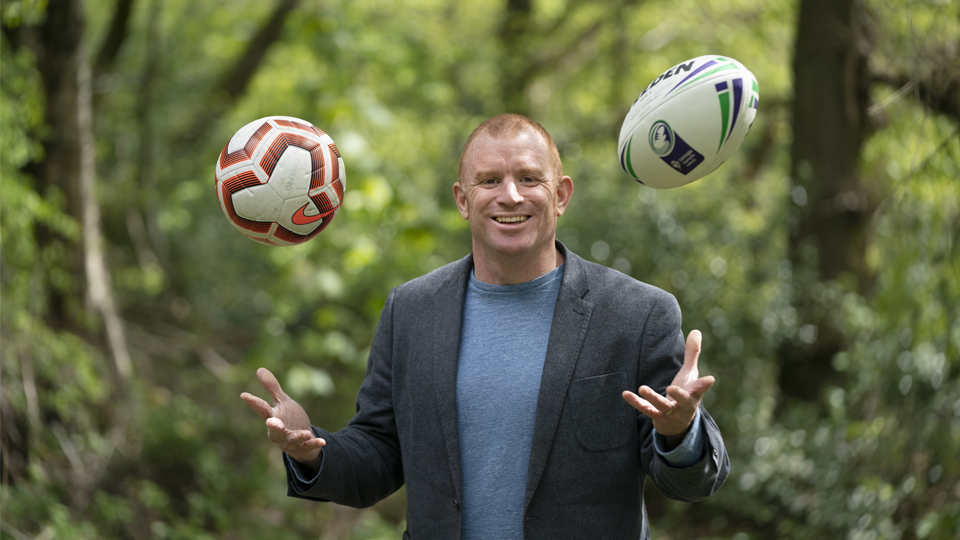 Chris Thair throwing a football and rugby ball in the air in front of trees.